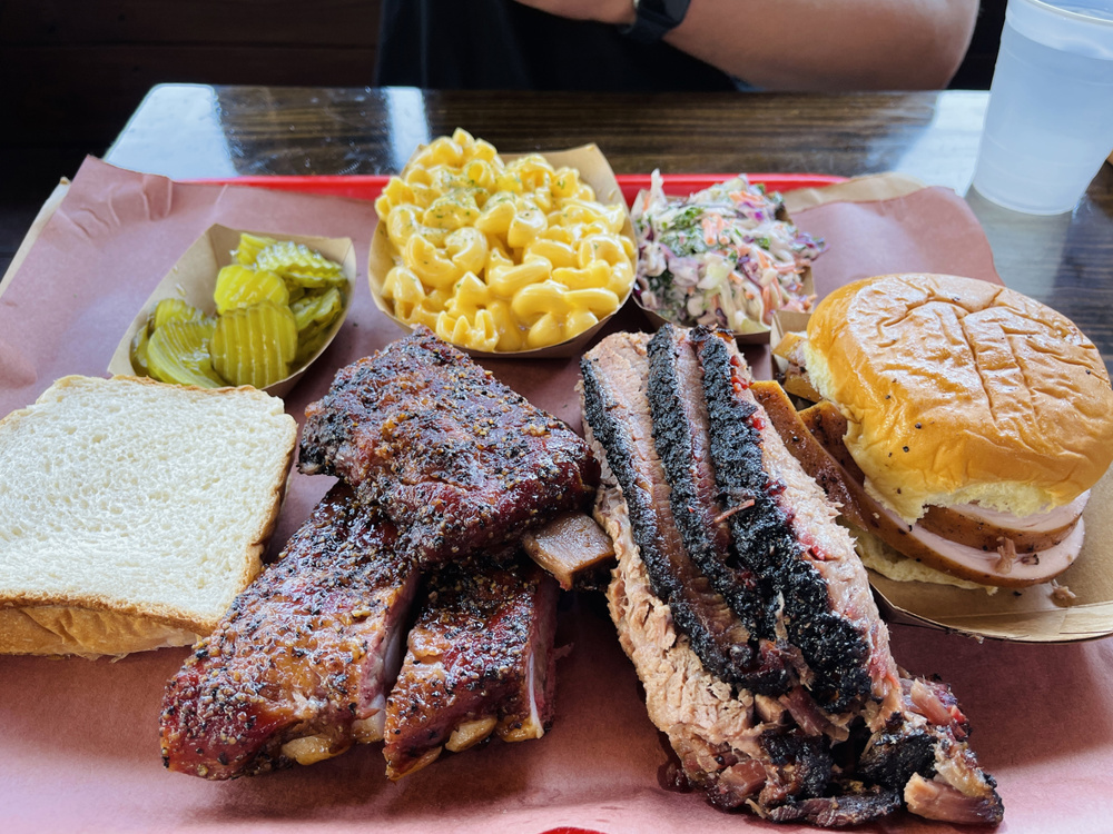 A large platter of barbecue meats and sides, including pork ribs, beef brisket, smoked turkey, mac and cheese, cole slaw, pickles, and bread
