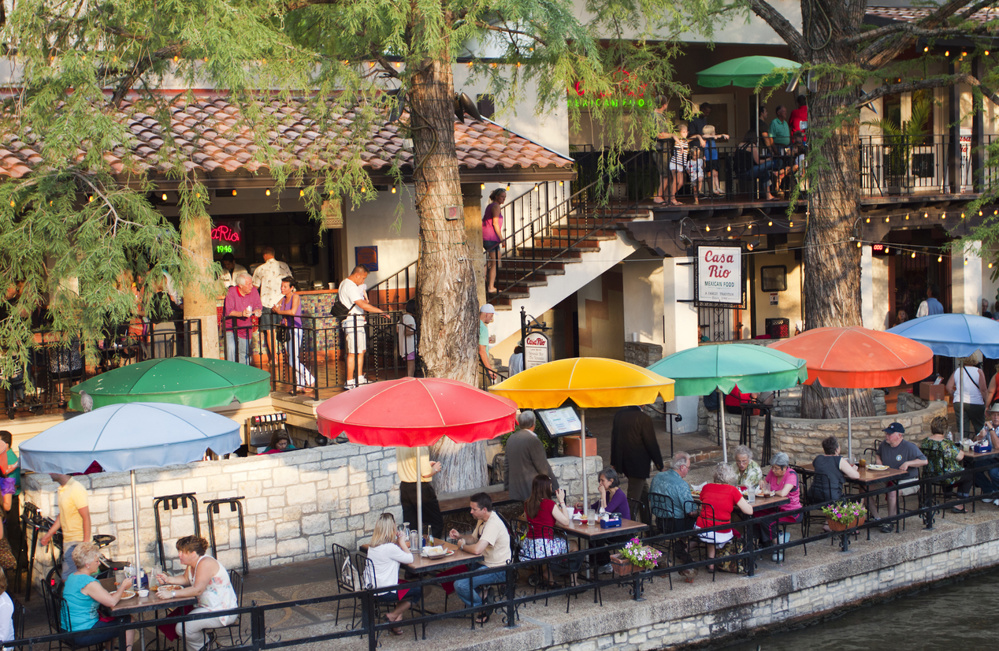 San Antonio River-walk scene with crowds of tourists dining under bright umbrellas, partying, and strolling along the river
